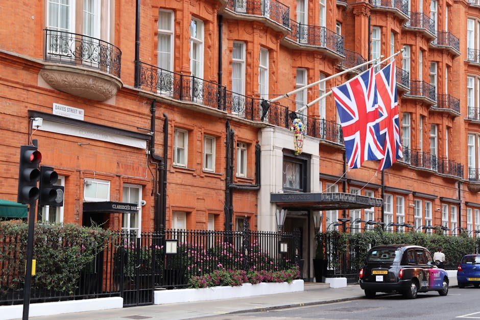 Exterior view of the Claridge's hotel in Berkeley Square, Mayfair, showing a red-brick façade with multiple windows and black wrought iron balconies. Two Union Jack flags are prominently displayed above the entrance, which is framed by a black awning with the hotel’s name. A black metal fence surrounds a landscaped area with pink flowering bushes along the sidewalk. A traffic light is visible on the left, and three cars are parked along the street. The scene is illuminated with natural daylight, highlighting the building’s classic architecture and tidy appearance, reflecting a well-maintained, high-end property, with no visible dirt or debris. The image emphasizes the upscale environment typical of Mayfair, complemented by the hotel's exterior cleanliness and maintained façade, aligning with the sophisticated area associated with premium surface cleaning and maintenance services, as promoted by Mayfair Carpet Cleaning.