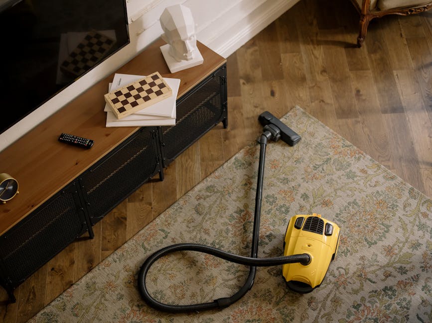 A woman performing surface cleaning and deep cleaning on a traditional patterned area rug using a vacuum cleaner in a living room. The rug features intricate floral motifs in shades of beige, green, and orange, and is situated on a light wooden floor. The woman is dressed casually in a beige jacket, blue jeans, and white sneakers, and is standing next to a brown leather sofa. The room is well-lit with natural light, and the overall cleanliness of the space suggests professional sanitisation and routine maintenance by Mayfair Carpet Cleaning, highlighting their domestic cleaning services for Berkeley Square flats.