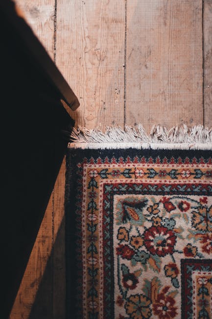 A detailed view of a multicoloured, patterned rug with intricate floral and geometric motifs lying on a polished wooden floor. The rug features a fringe edge at the bottom, with slightly frayed white tassels. To the left, part of a dark wooden piece of furniture is visible, casting a shadow over part of the rug. The room is illuminated with soft, natural lighting, highlighting the texture and vibrant colors of the carpet. The scene reflects a clean and well-maintained domestic space, showcasing surface cleaning and care for antique rugs, aligned with services offered by Mayfair Carpet Cleaning.