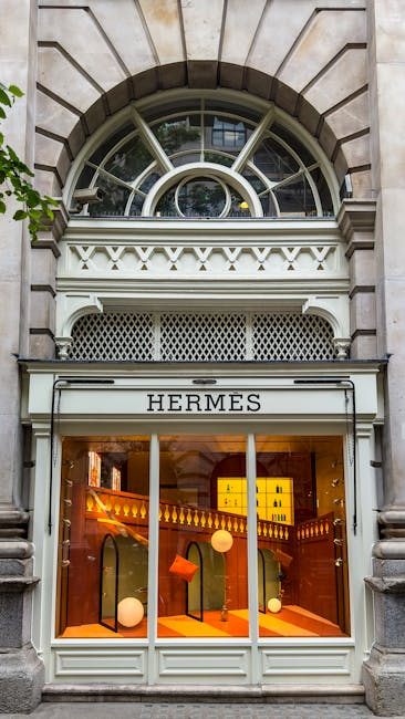 Photograph of the exterior of a building with a curved, semi-circular window above a retail store. The façade features a stone archway surrounding the window, with decorative elements and white trim. The store, named 'HERMES,' has large glass display windows showcasing illuminated interior fixtures, including soft orange lighting, spherical display props, and shelves with neatly arranged products. The scene is well-lit, highlighting the clean, polished surface of the storefront and the architectural details of the stonework and window framing. The overall appearance emphasizes a high-end retail environment in a sophisticated urban setting, aligning with concepts of surface cleaning and maintenance as part of professional hygiene and presentation, consistent with services provided by Mayfair Carpet Cleaning in their focus on deep cleaning and sanitisation.
