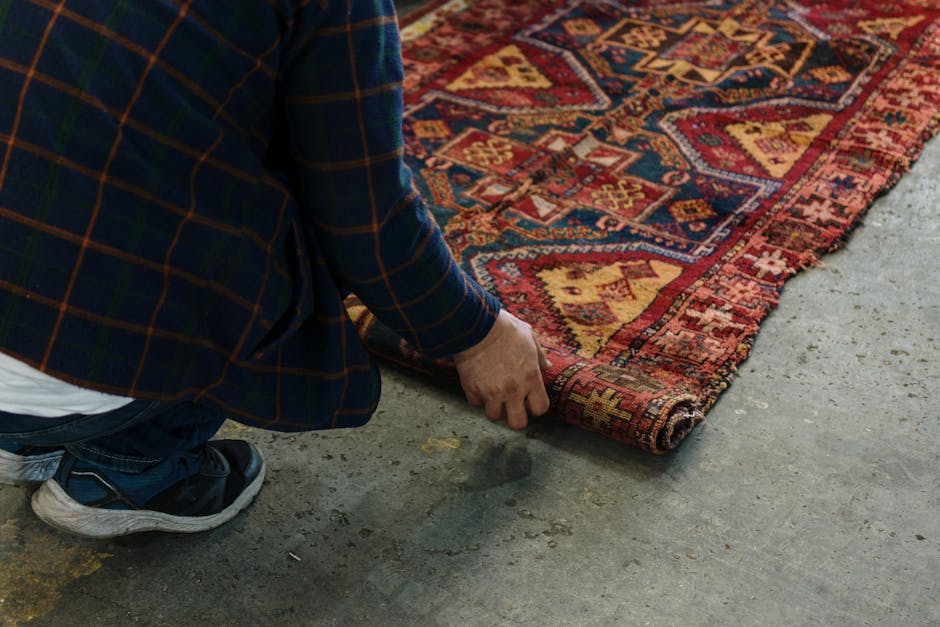 A detailed view of a multicoloured, patterned rug with intricate floral and geometric motifs lying on a polished wooden floor. The rug features a fringe edge at the bottom, with slightly frayed white tassels. To the left, part of a dark wooden piece of furniture is visible, casting a shadow over part of the rug. The room is illuminated with soft, natural lighting, highlighting the texture and vibrant colors of the carpet. The scene reflects a clean and well-maintained domestic space, showcasing surface cleaning and care for antique rugs, aligned with services offered by Mayfair Carpet Cleaning.