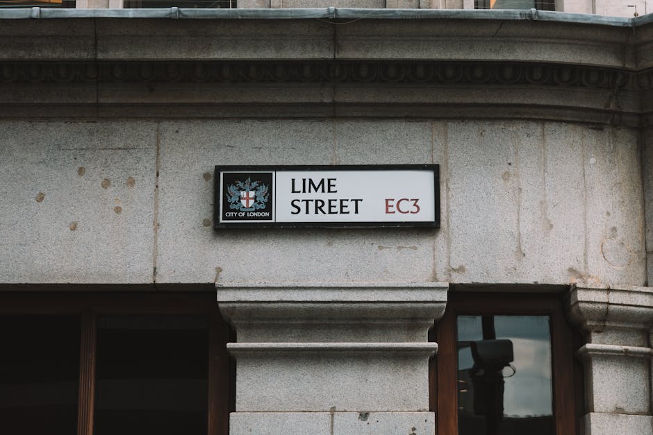 Close-up of a street sign mounted on a beige stone building at the corner of Lime Street, City of London, with black lettering on a white background and red indicating the postal district EC3. The sign features the City of London crest, which includes a shield with a red cross and sword, topped with a crown, and additional insignia. The building's surface displays a smooth, clean stone finish with visible joints and some weathering marks. Surrounding the sign, part of a window with a wooden frame and a reflective glass pane is visible beneath the sign, and the photo is taken outdoors in natural light, capturing the urban environment of central London.
