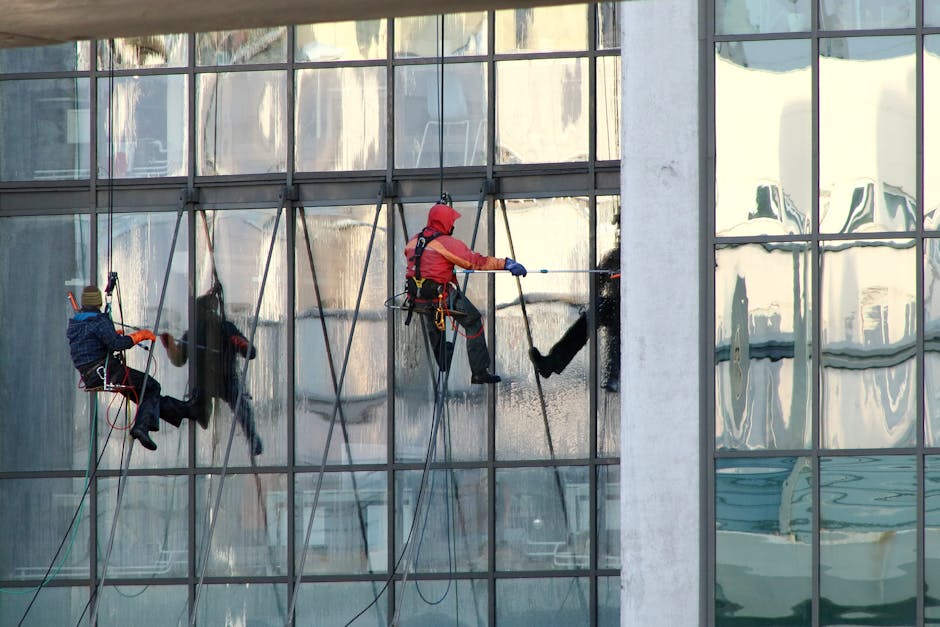 Two professional window cleaners from Mayfair Carpet Cleaning are suspended on ropes, performing surface cleaning and deep cleaning of large, reflective glass windows on a high-rise commercial building. One cleaner, dressed in a red jacket and helmet, is using a spray and squeegee to thoroughly clean the glass surface, creating a streak-free, shiny finish. The other cleaner, wearing a dark jacket and helmet, is positioned on a separate rope, holding cleaning tools and working on an adjacent window pane. The building's exterior features multiple glass panels with a sleek, modern design, reflecting the urban environment and surrounding buildings. The scene is well-lit by natural daylight, emphasizing the clarity and cleanliness of the glass surfaces. This image illustrates professional sanitisation and surface cleaning techniques used in commercial window maintenance, highlighting the expertise of Mayfair Carpet Cleaning in high-level cleaning services.