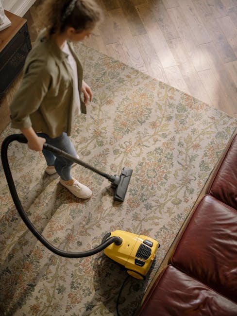 A woman using a yellow canister vacuum cleaner to deep clean a patterned area rug with floral motifs in a living room. She is wearing a beige jacket, blue jeans, and white sneakers, and is standing on the wooden floor adjacent to the rug. The room features natural lighting, with a red leather sofa partially visible at the edge of the frame. The surface cleaning process highlights the attention to hygiene and maintenance, with the rug appearing plush and clean. The image exemplifies domestic cleaning, with Mayfair Carpet Cleaning providing professional carpet and surface cleaning services to ensure a pristine living environment.