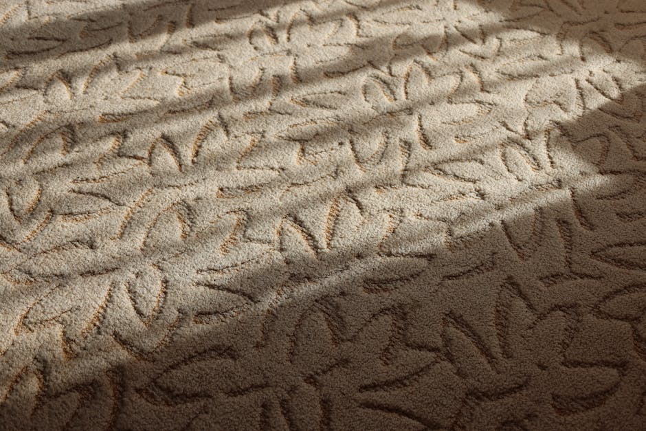 A woman using a yellow canister vacuum cleaner to deep clean a patterned area rug with floral motifs in a living room. She is wearing a beige jacket, blue jeans, and white sneakers, and is standing on the wooden floor adjacent to the rug. The room features natural lighting, with a red leather sofa partially visible at the edge of the frame. The surface cleaning process highlights the attention to hygiene and maintenance, with the rug appearing plush and clean. The image exemplifies domestic cleaning, with Mayfair Carpet Cleaning providing professional carpet and surface cleaning services to ensure a pristine living environment.