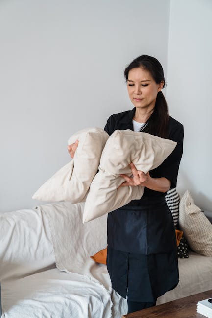 A professional cleaner, a woman with dark hair pulled back, dressed in a black uniform, is standing in a bright living room holding a beige pillow and a folded blanket. Behind her is a white sofa covered with a light-colored throw, and several cushions in neutral tones, including beige and black-and-white patterns. The room features white walls and a wooden side table. The scene is well-lit with natural light, emphasizing the clean and tidy appearance of the space, indicating surface and textile cleaning associated with Mayfair Carpet Cleaning's upholstery cleaning services in Grosvenor Square.