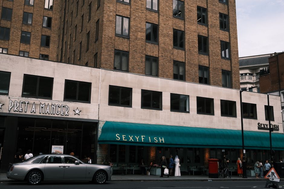 Image of a multi-story building with a brick facade and several dark, reflective window panes. The ground floor features a restaurant with a white section and a large teal awning displaying the name 'SEXYFISH' in white lettering. To the left of the restaurant is another establishment with a sign that reads