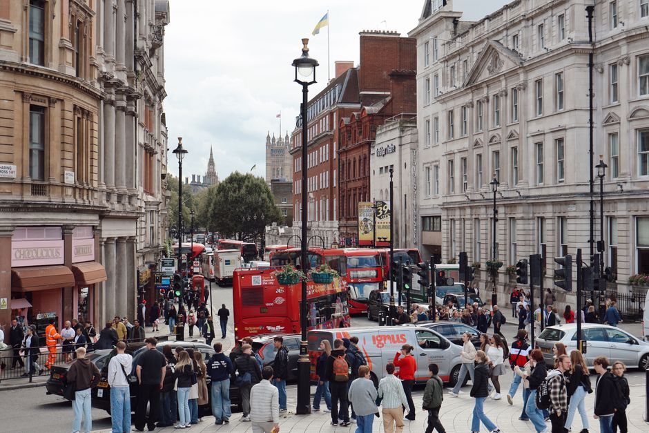 A busy street scene in Mayfair, London, featuring a mix of classic and modern architecture with white and red brick facades. The street is filled with pedestrians walking on the pavement, some waiting to cross at a traffic light, while others are engaged in conversation or looking at their phones. Multiple vehicles, including red double-decker buses, cars, and a white van labeled 'Ignus', occupy the road, with some stopped at the traffic signals. Lampposts line the sidewalks, and greenery in the form of trees adds contrast to the urban setting. The weather appears overcast, with diffuse lighting creating soft shadows, and the background showcases iconic London landmarks like the Elizabeth Tower partially visible among the buildings. This scene exemplifies typical city life in Mayfair, highlighting the bustling environment and diverse activity.