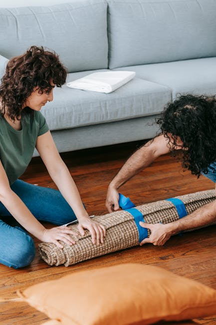 Two individuals are engaged in surface cleaning by rolling out a beige carpet with blue straps on a polished wooden floor in a living room. One person, a woman with curly brown hair wearing a green top and blue jeans, is holding and positioning the carpet, while the other person, a man with curly dark hair in a blue shirt, is assisting by pulling or securing it. The background features a light grey fabric sofa with a white cushion, and the room is well-lit with natural light. The scene emphasizes domestic cleaning and maintenance, with a focus on preparing the carpet for further cleaning or placement, showcasing attention to hygiene and surface care in a home environment. Mayfair Carpet Cleaning offers expert services for effective deep cleaning and sanitisation of carpets and surfaces.