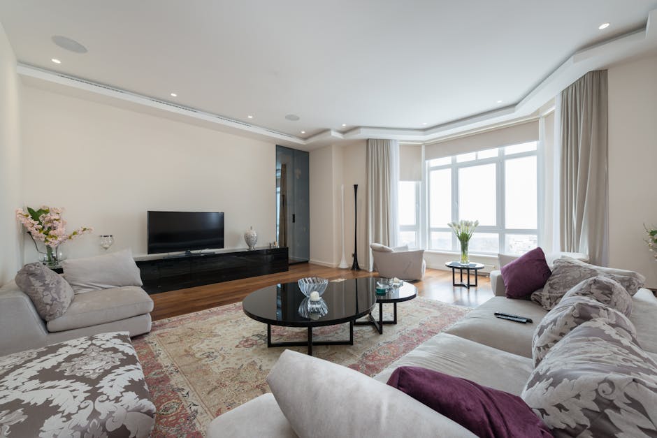 A bright and spacious living room in a Mayfair flat featuring a large window with white curtains allowing natural daylight to illuminate the space. The room has a white ceiling with recessed lighting and plain white walls. The flooring is polished hardwood. There are two cream-colored sofas with patterned and solid purple cushions, arranged around a round black coffee table with decorative glass items on top. Against the wall, a black media console supports a flat-screen TV and decorative vases. The room is neatly maintained with no visible dust or clutter, exemplifying professional domestic cleaning. A tall, slender lamp stands near the window, and fresh flowers in vases add a touch of elegance. Mayfair Carpet Cleaning specializes in surface cleaning and deep cleaning services to ensure homes remain hygienic and well-maintained.