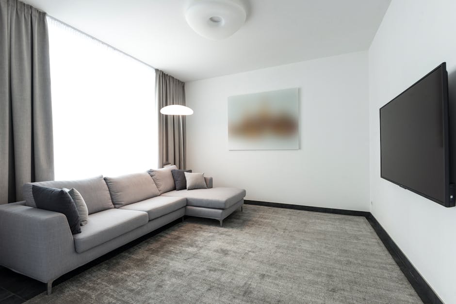 A modern living room with a light grey fabric sofa placed against a white wall, decorated with matching cushions; a large window with grey curtains allowing natural light to fill the space; a white ceiling with a simple circular light fixture; a flat-screen television mounted on the right wall; the room features a neutral-toned carpet and a piece of abstract art on the wall. The clean and tidy appearance suggests recent surface cleaning and maintenance, reflecting the professional domestic cleaning services offered by Mayfair Carpet Cleaning, including deep cleaning and sanitisation for residential spaces.