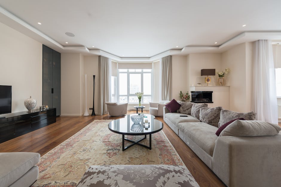 A bright and spacious living room in a Mayfair flat featuring a large window with white curtains allowing natural daylight to illuminate the space. The room has a white ceiling with recessed lighting and plain white walls. The flooring is polished hardwood. There are two cream-colored sofas with patterned and solid purple cushions, arranged around a round black coffee table with decorative glass items on top. Against the wall, a black media console supports a flat-screen TV and decorative vases. The room is neatly maintained with no visible dust or clutter, exemplifying professional domestic cleaning. A tall, slender lamp stands near the window, and fresh flowers in vases add a touch of elegance. Mayfair Carpet Cleaning specializes in surface cleaning and deep cleaning services to ensure homes remain hygienic and well-maintained.