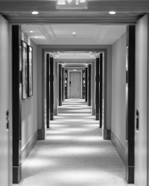 Black and white image of a long, symmetrical corridor in a commercial or residential building, featuring wooden door frames on both sides, evenly spaced along the hallway. The walls are smooth and decorated with framed pictures, creating a tidy and professional appearance. The ceiling includes recessed lighting fixtures that illuminate the pathway, enhancing the sense of cleanliness and order. The polished wooden flooring appears well-maintained and free of dust or debris, reflecting the ambient light to emphasize the surface's shine. Mayfair Carpet Cleaning specializes in surface cleaning, deep cleaning, and sanitisation, ensuring hygienic and spotless environments in both residential and commercial settings. This image showcases a pristine hallway space, highlighting the importance of regular cleaning maintenance as part of a comprehensive checklist for buildings in Mayfair, W1K.