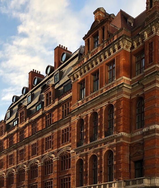 A close-up view of an ornate, historic red brick building with multiple stories, featuring arched windows with decorative stone lintels and protruding bay windows. The roof has dormer windows with rounded tops and metal cladding, with chimneys and vent pipes visible on top. The building's facade shows detailed stonework and cornices, and the sunlight highlights the texture of the brickwork. The sky above is partly cloudy, providing natural lighting that accentuates the architectural details. This setting emphasizes traditional exterior maintenance, aligning with Mayfair Carpet Cleaning's focus on surface cleaning and property preservation for residential and commercial buildings.
